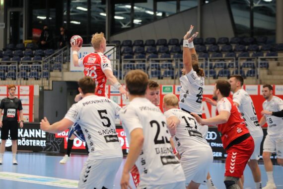 Ein Handballspieler in einem roten Trikot springt, um den Ball zu werfen, während mehrere Spieler in weißen Trikots versuchen, ihn während eines Hallenspiels zu blockieren. Im Hintergrund sind Zuschauer und leere Sitze zu sehen.