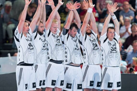 Sechs männliche Handballspieler in passenden weißen und schwarzen Trikots stehen mit erhobenen Armen in einer Reihe und blockieren gemeinsam. Im Hintergrund sieht eine Gruppe von Zuschauern zu.