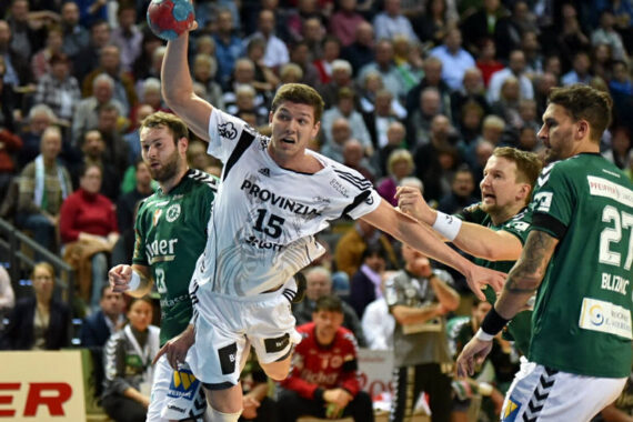 Ein Handballspieler in einem weißen Trikot springt, um den Ball zu schießen, während drei gegnerische Spieler in Grün versuchen, ihn zu blockieren. Das Spiel ist intensiv, und im Hintergrund sieht eine Menge Zuschauer von der Tribüne aus zu.