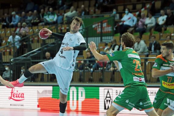 Ein Handballspieler in einem weißen Trikot springt, um den Ball zu werfen, während zwei gegnerische Spieler in grünen Trikots mit orangefarbenen Streifen versuchen, ihn während eines Spiels in einer überfüllten Halle zu verteidigen.