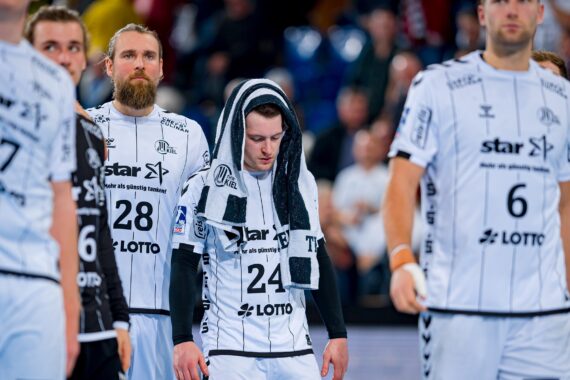 Eine Gruppe männlicher Handballspieler in weißen Trikots steht auf dem Spielfeld; ein Spieler in der Mitte, die Nummer 24, blickt mit einem Handtuch über dem Kopf nach unten, was auf Enttäuschung oder Erschöpfung schließen lässt.