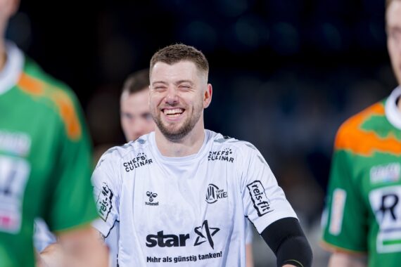 Ein Mann in einem weißen Sporttrikot lächelt breit während einer Hallensportveranstaltung und steht zwischen zwei anderen Spielern in grünen Trikots. Der Hintergrund ist unscharf, um die Aufmerksamkeit auf seinen freudigen Ausdruck zu lenken.