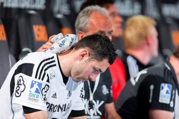 Ein männlicher Sportler in einem schwarz-weißen Trikot sitzt auf einer Bank und schaut nach unten, während die Hand eines Mannschaftskameraden unterstützend auf seiner Schulter ruht. Andere Mannschaftskameraden sitzen in der Nähe, verschwommen im Hintergrund.