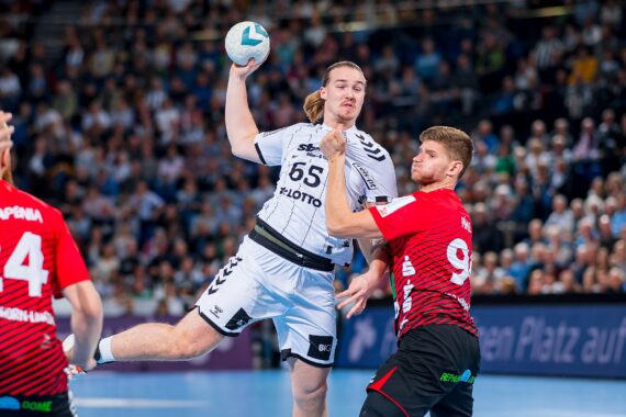 Ein Handballspieler in einem weißen Trikot springt, um den Ball zu werfen, während ein Spieler in einem rot-schwarzen Trikot versucht, ihn zu blockieren. Das Spiel findet auf einem Hallenplatz statt, während im Hintergrund eine Menschenmenge zuschaut.