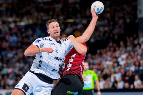 Zwei männliche Handballspieler liefern sich einen intensiven Wettkampf; der eine in einem weißen Trikot versucht, den Ball zu schießen oder zu passen, während ein Spieler in einem rot-schwarzen Trikot versucht, ihn zu blockieren. Im Hintergrund sind eine unscharfe Menge und ein Schiedsrichter zu sehen.