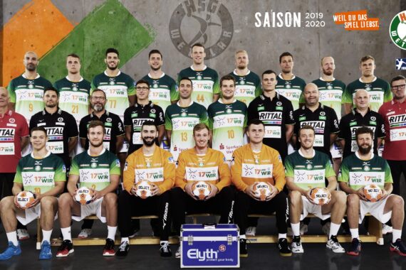 Eine Herren-Handballmannschaft stellt sich in drei Reihen in grün-weißen Trikots für ein Gruppenfoto auf. Vier Spieler in Orange sitzen vorne und halten Bälle. Die Mitarbeiter der Mannschaft stehen auf jeder Seite, und im Hintergrund sind Logos und Saisondaten zu sehen.