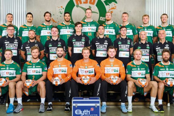 Eine Männer-Handballmannschaft in grünen Trikots posiert für ein Gruppenfoto in einer Halle. Trainer und Betreuer stehen hinter den Spielern, die vorne sitzen; zwei Spieler in orangefarbenen Trikots halten Handbälle. Im Hintergrund sind ein Mannschaftslogo und ein Sponsor zu sehen.