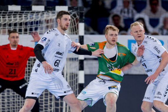 Drei Handballspieler treten in der Nähe des Tores gegeneinander an, wobei zwei Spieler in Weiß gegen einen Spieler in Grün verteidigen, während ein Torwart in einem roten Trikot im Hintergrund auf der Hut ist.