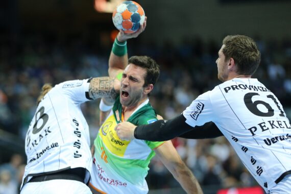 Ein Handballspieler in einem grün-gelben Trikot springt mit dem Ball, während zwei gegnerische Spieler in weißen Trikots versuchen, ihn während eines Spiels zu blockieren. Die Szene ist intensiv und actionreich.