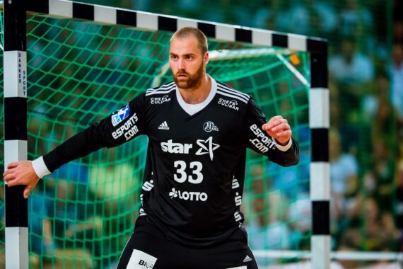 Ein Handballtorwart in einem schwarzen Trikot mit der Nummer 33 steht aufmerksam vor dem Tor, bereit zur Verteidigung. Das Netz und der Torpfosten sind hinter ihm zu sehen, und die Zuschauer sind im Hintergrund verschwommen.