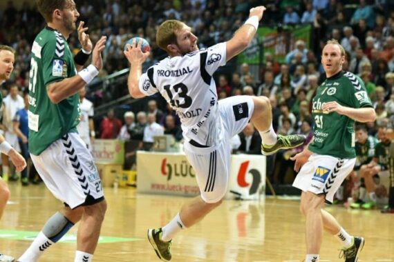 Ein Handballspieler in einem weißen Trikot springt, um den Ball zu schießen, während zwei Verteidiger in grünen Trikots versuchen, ihn auf einem Hallenplatz zu blockieren, während im Hintergrund eine Menschenmenge zusieht.