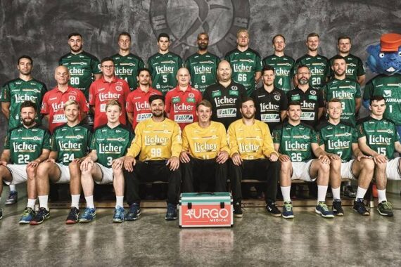 Eine Herren-Handballmannschaft in grünen Trikots posiert für ein Gruppenfoto in drei Reihen, mit drei Personen in gelben und roten Trikots in der ersten Reihe. Ganz rechts steht ein blaues Maskottchen.