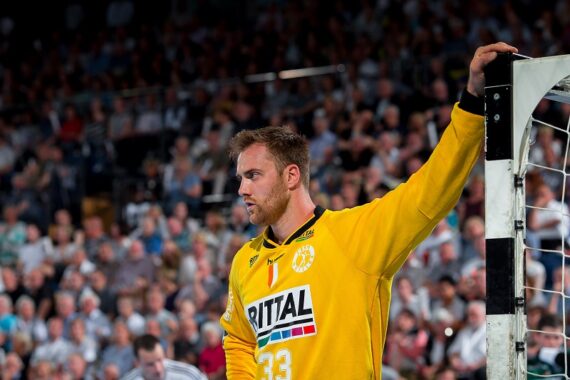 Ein männlicher Handballtorwart in einem gelben Trikot mit der Nummer 33 steht am Torpfosten und schaut konzentriert. Der Hintergrund zeigt eine unscharfe Zuschauermenge in einer Halle.