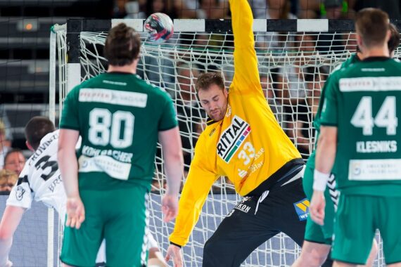 Ein Handballtorwart in einem gelben Trikot streckt sich aus, um einen Schuss abzublocken, während drei Spieler in grünen Trikots während eines Spiels in der Nähe des Tores stehen.