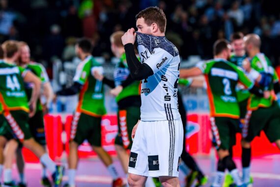 Ein Handballspieler in einem weiß-schwarzen Trikot bedeckt sein Gesicht mit seinem Trikot, während Spieler in grünen und orangefarbenen Trikots im Hintergrund auf einem hell erleuchteten Hallenplatz feiern.