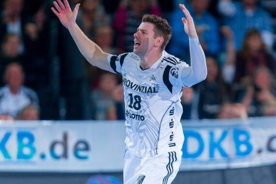 Ein männlicher Handballspieler in einem weiß-schwarzen Trikot mit der Nummer 18 reißt die Arme hoch und jubelt auf einem Hallenplatz, im Hintergrund sind Zuschauer und ein DKB.de-Banner zu sehen.