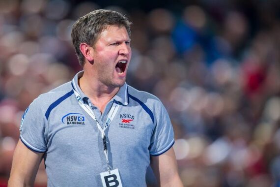 Ein Mann in einem grauen Polohemd mit Handball-Logos schreit, wahrscheinlich als Trainer während eines Spiels, mit einer unscharfen Menschenmenge im Hintergrund. Er trägt ein Schlüsselband mit einem Abzeichen mit der Aufschrift B.