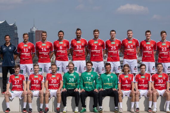 Eine Herren-Handballmannschaft posiert im Freien. Die Spieler in roten Trikots stehen in zwei Reihen, vor ihnen sitzen drei Torhüter in Grün. Trainer und Betreuer in dunkler Kleidung stehen an beiden Enden. Im Hintergrund sind eine Stadtlandschaft und der blaue Himmel zu sehen.