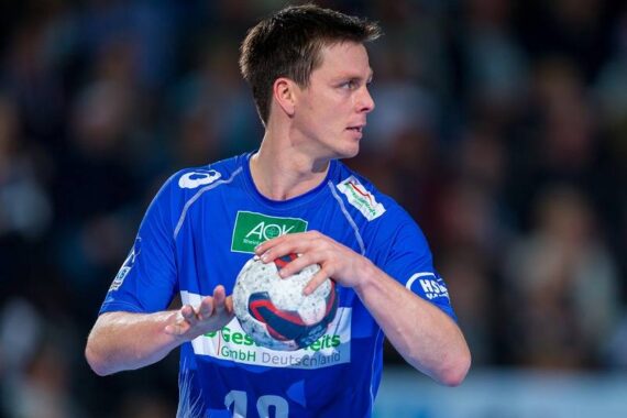 Ein männlicher Handballspieler in einem blauen Trikot hält einen Ball und schaut nach links, als ob er sich auf den Wurf vorbereitet. Der Hintergrund ist mit Zuschauern unscharf.