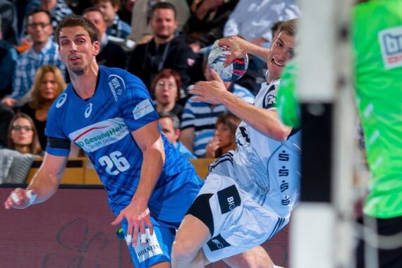 Zwei männliche Handballspieler in blau-weißen Trikots kämpfen während eines Spiels um den Ball, während im Hintergrund unscharfe Zuschauer zuschauen und im Vordergrund ein Schiedsrichter in einem grünen Hemd zu sehen ist.