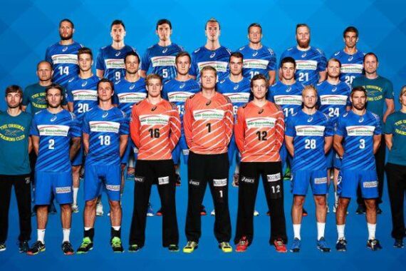 Eine Herren-Handballmannschaft posiert in drei Reihen vor einem blauen Hintergrund. Die Spieler tragen blau-weiße Trikots, während drei Spieler in der vorderen Reihe orangefarbene Trikots tragen. Die Trainer oder Betreuer in passenden blauen Hemden stehen an den Seiten.