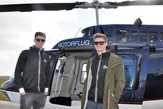 Zwei Männer mit Sonnenbrillen und Jacken stehen an einem bewölkten Tag vor einem Hubschrauber mit der Aufschrift ROTORFLUG, dessen Seitentür geöffnet ist.