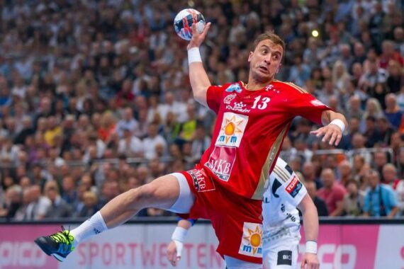 Ein männlicher Handballspieler in einem roten Trikot springt in der Luft, um den Ball während eines Spiels zu werfen. Im Hintergrund sind unscharfe Zuschauer und gegnerische Spieler in weißen Trikots zu sehen.