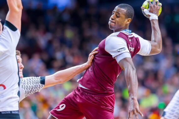 Ein Handballspieler in einem kastanienbraunen Trikot springt in der Luft und bereitet sich auf den Wurf vor, während ein gegnerischer Spieler in Weiß versucht, ihn während eines Spiels zu blockieren. Das Publikum ist unscharf im Hintergrund zu sehen.