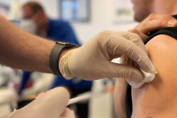 Nahaufnahme einer medizinischen Fachkraft mit Handschuhen, die den Oberarm einer Person mit einem Wattebausch reinigt, bevor sie eine Injektion gibt. Eine andere Person ist im Hintergrund unscharf zu erkennen.