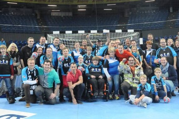 Eine große Gruppe von Menschen, darunter Kinder und Erwachsene, posieren gemeinsam auf einem Hallensportplatz vor einem Handballtor, viele tragen passende Sporttrikots und halten Handbälle und lächeln in die Kamera.