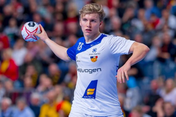 Ein männlicher Handballspieler in einem weiß-blauen Trikot bereitet sich darauf vor, den Ball während eines Spiels zu werfen, mit einer unscharfen Menschenmenge im Hintergrund.