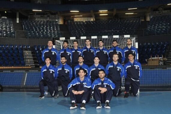 Eine Herren-Handballmannschaft in passenden blauen und schwarzen Trainingsanzügen posiert für ein Gruppenfoto vor einem Tor in einer Sporthalle mit leeren Sitzen im Hintergrund.