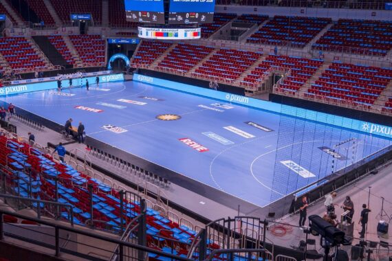 Eine große Sporthalle mit einem blauen Handballfeld, umgeben von roten und blauen Sitzen. In der Nähe des Spielfelds befinden sich einige Personen und Geräte, und digitale Werbetafeln säumen den Rand. Die Halle ist größtenteils leer.