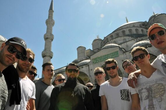 Eine Gruppe junger Männer mit Sonnenbrillen posiert an einem sonnigen Tag für ein Foto vor der Blauen Moschee in Istanbul, Türkei. Die Kuppeln und das Minarett der Moschee sind im Hintergrund zu sehen.