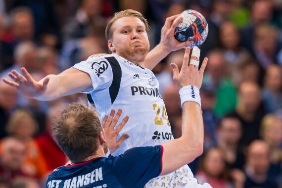 Zwei männliche Handballspieler in Aktion während eines Spiels. Der Spieler in Weiß springt mit dem Ball, während er von einem Spieler in Dunkelblau verteidigt wird. Der Hintergrund zeigt eine unscharfe Zuschauermenge.