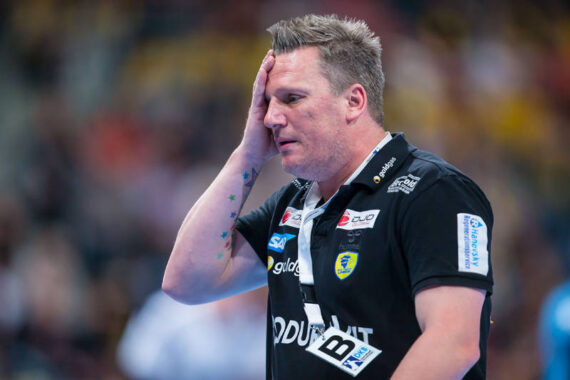 Ein männlicher Handballtrainer, der ein schwarzes Hemd mit Sponsorenlogos trägt, steht auf dem Spielfeld und berührt während eines Spiels mit besorgter oder gestresster Miene seine Stirn.
