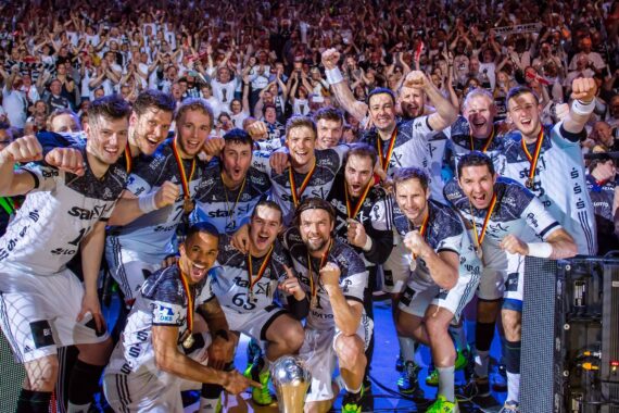 Ein jubelndes Männer-Handballteam posiert lächelnd und jubelnd mit Medaillen um den Hals und einem Pokal vor sich, während im Hintergrund eine große, begeisterte Menge feiert.