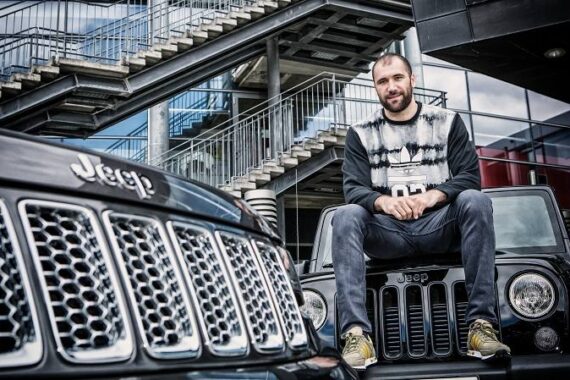 Ein Mann sitzt auf der Motorhaube eines schwarzen Jeeps, im Vordergrund steht ein weiterer Jeep. Im Hintergrund sind Metalltreppen und moderne Architektur zu sehen.