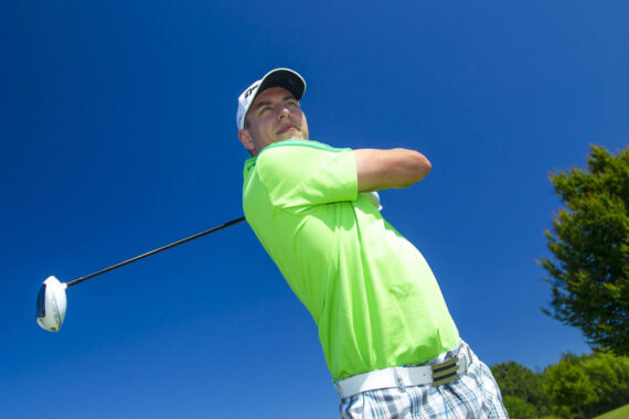 Ein Golfer in einem hellgrünen Hemd und einer weißen Kappe führt einen Schwung aus und hält einen Golfschläger über die Schulter. Der Himmel ist blau und die Bäume im Hintergrund sind klar.