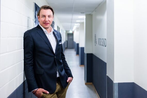 Ein Mann in dunkler Anzugjacke und khakifarbener Hose steht in einem hellen Flur, lehnt lässig an einer weiß-blauen Wand, hat die Hände in den Taschen und schaut leicht lächelnd in die Kamera.
