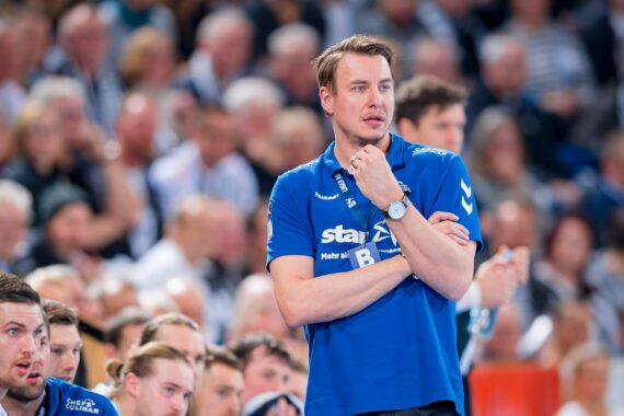 Ein Mann in einem blauen Sporthemd steht mit verschränkten Armen und einem nachdenklichen Gesichtsausdruck bei einer Sportveranstaltung, während im Hintergrund Zuschauer und Mannschaftsmitglieder zu sehen sind.