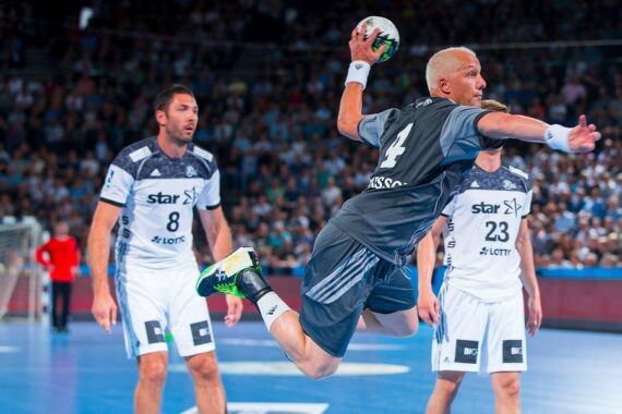 Ein Handballspieler in einem grauen Trikot springt mit einem Ball in die Luft und bereitet sich auf einen Schuss vor, während zwei Verteidiger in weißen Trikots während eines Spiels in einer überfüllten Arena zusehen.