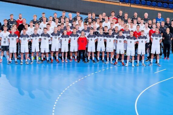 Eine große Gruppe von männlichen Sportlern und Trainern posiert gemeinsam auf einem Hallenhandballfeld. Sie tragen passende Sportuniformen in Weiß und Schwarz mit Nummern, einige tragen rote Torwarttrikots.