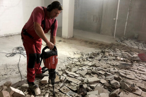 Eine Person mit Gehörschutz benutzt einen Presslufthammer, um Fliesen und Beton auf dem Boden eines großen, staubigen, unfertigen Raums zu zertrümmern. Trümmer und Scherben liegen auf dem Boden verstreut.