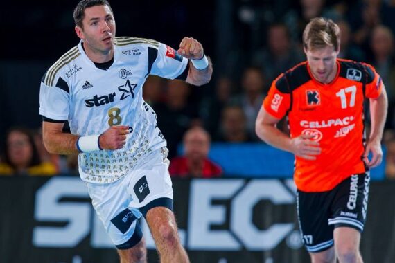 Zwei männliche Handballspieler in Aktion während eines Spiels; einer in einem weißen Trikot mit der Nummer 8 und der andere in einem orange-schwarzen Trikot mit der Nummer 17, die auf einem Spielfeld mit unscharfen Zuschauern im Hintergrund laufen.