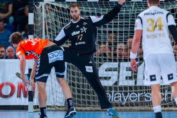 Ein schwarz gekleideter Handballtorwart bereitet sich darauf vor, einen Schuss abzublocken, indem er seine Arme und Beine vor dem Tor ausbreitet. Zwei gegnerische Spieler in orange-weißen Trikots stehen in der Nähe auf dem Spielfeld.