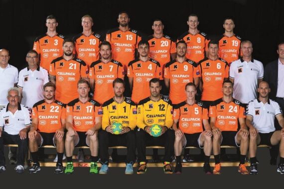 Eine Handballmannschaft der Männer posiert in einer Halle für ein Gruppenfoto in orange-schwarzen Trikots. Trainer und Betreuer in weißer Kleidung sitzen und stehen auf beiden Seiten; zwei Torhüter halten Handbälle vor sich.