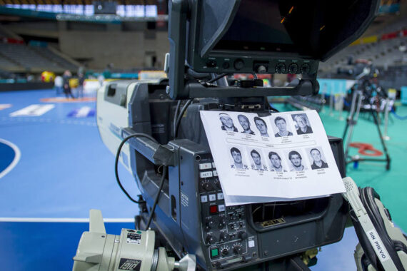 Nahaufnahme einer Fernsehkamera neben einer Sporthalle, mit einem Blatt mit gedruckten Fotos und Namen von Personen, die an die Kamera geklebt sind, und unscharfen Figuren und Geräten im Hintergrund.
