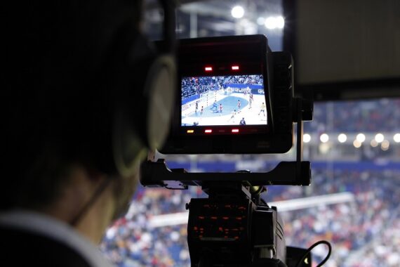 Nahaufnahme einer Fernsehkamera, die eine Sportveranstaltung in einer Halle filmt, wobei das Spiel und die Spieler auf dem Monitor der Kamera zu sehen sind und die Zuschauer im Hintergrund verschwimmen.