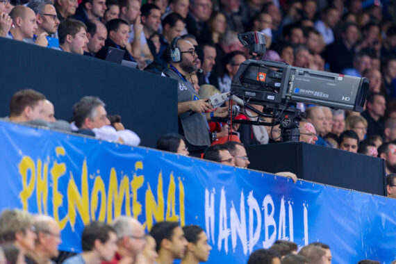 Ein Kameramann filmt ein Handballspiel von der Tribüne aus, umgeben von einer Menge von Zuschauern. Vor ihnen hängt ein blaues Banner mit der Aufschrift PHENOMENAL HANDBALL! in gelben Buchstaben.